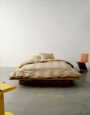 Bett mit beiger Bettwäsche auf einem Holzrahmen, frontal fotografiert, mit modernem gelbem und hölzernem Stuhl im Hintergrund.