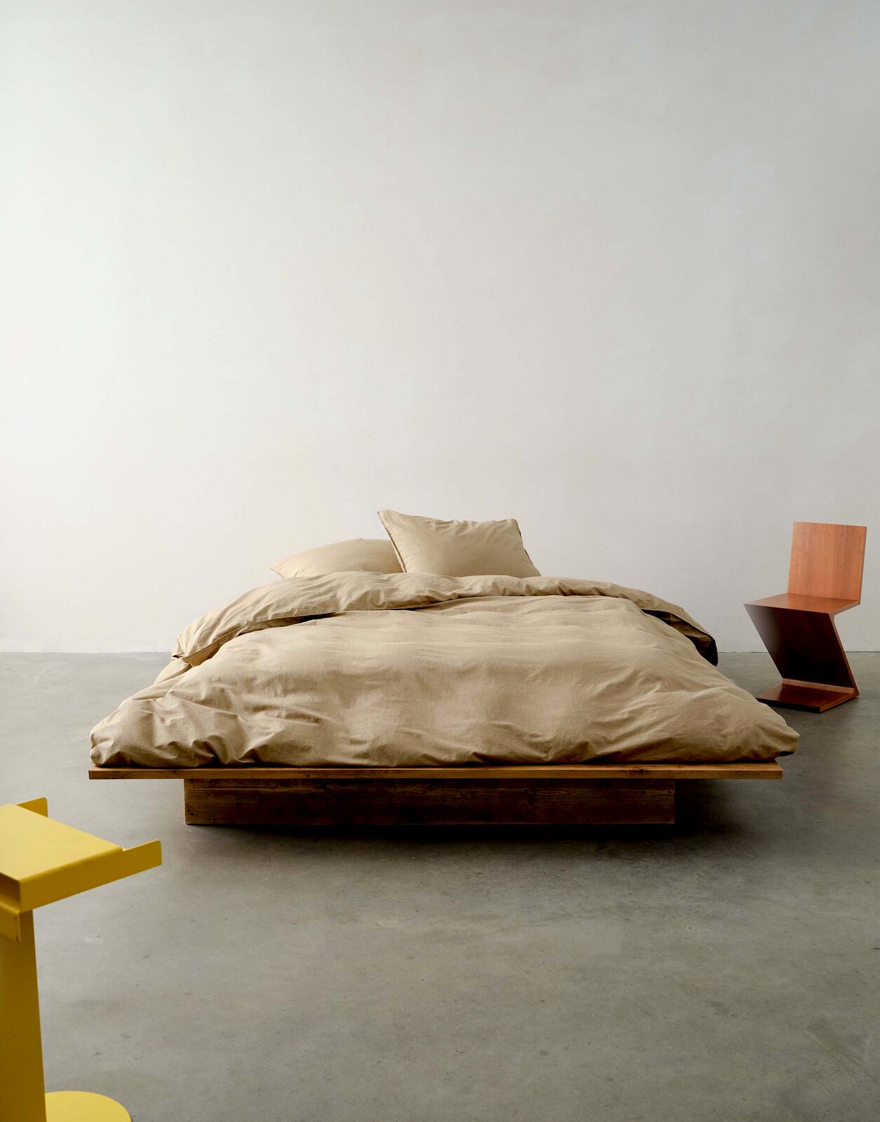 Bett mit beiger Bettwäsche auf einem Holzrahmen, frontal fotografiert, mit modernem gelbem und hölzernem Stuhl im Hintergrund.