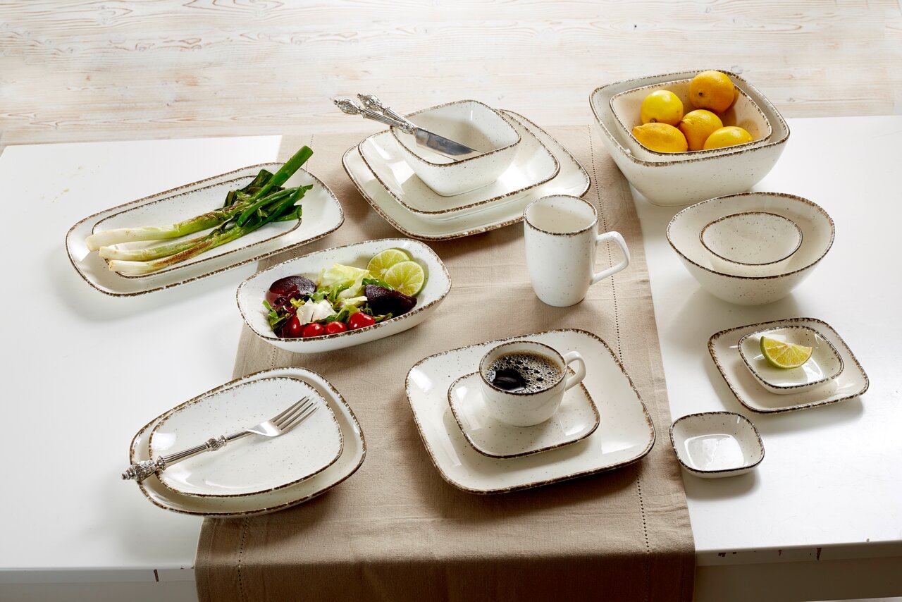 Vogelperspektive auf ein Set von beigen Keramikgeschirr mit rustikalem Design, darunter eine Dipschale, Teller, Tassen und Schalen, dekoriert mit Lebensmitteln und Besteck.