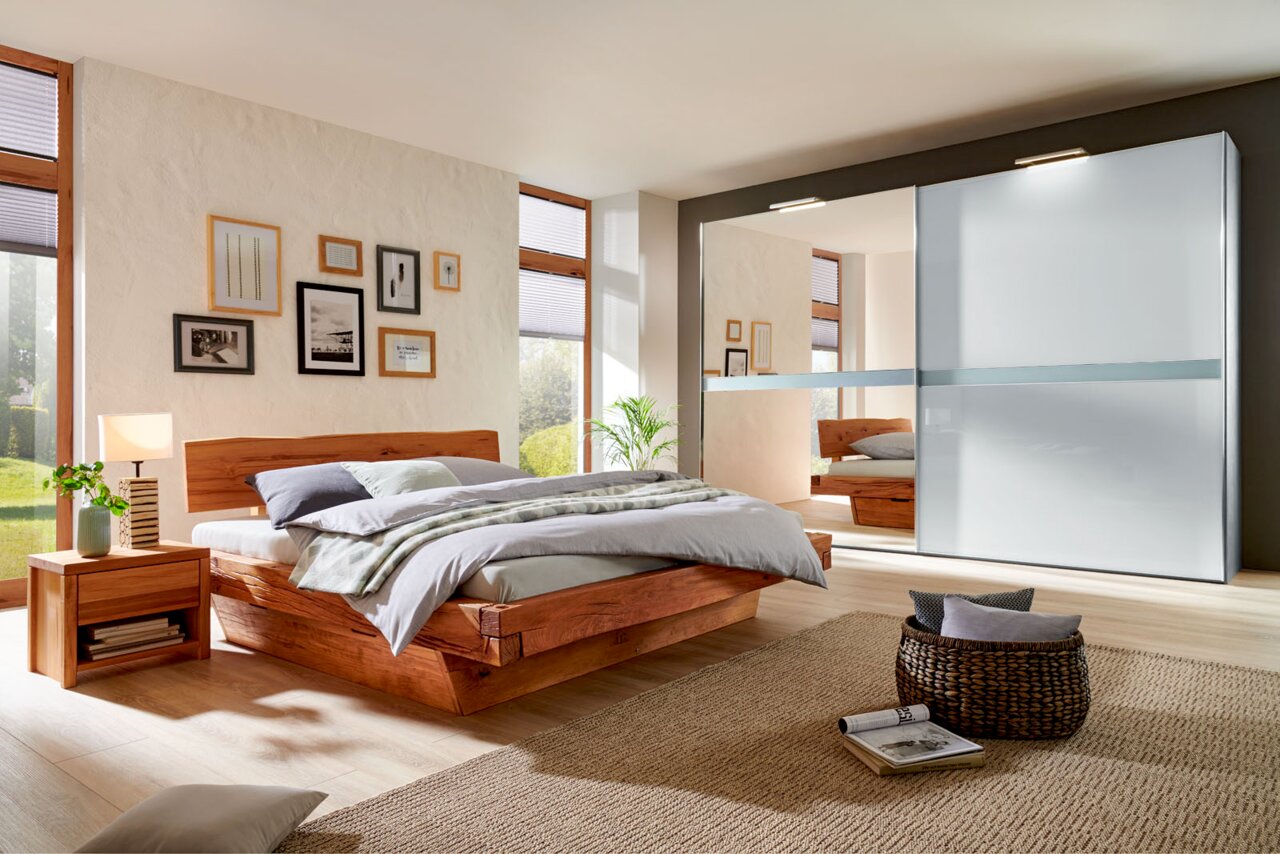 Linea Natura Bed AMAZON Modernes Schlafzimmer mit einem eleganten Holzbett und einem großen Kleiderschrank mit Schiebetüren, seitliche Perspektive.