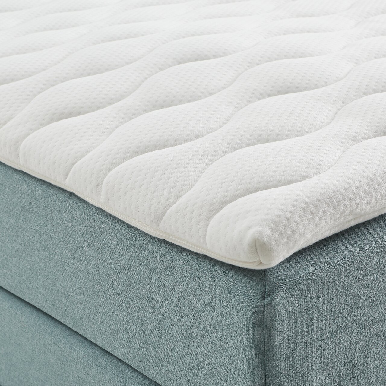 Nahaufnahme eines Boxspringbetts mit weißem, gestepptem Topper und hellblauem Stoffbezug, seitliche Perspektive