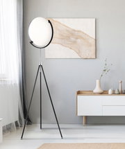 Moderne LED-Stehleuchte mit rundem, weißem Lampenschirm auf einem dreibeinigen schwarzen Stativ, seitliche Perspektive im Wohnzimmer.