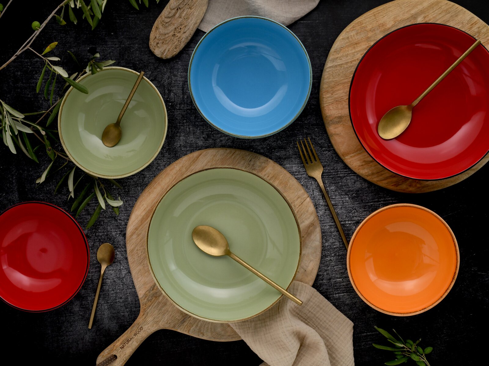 Draufsicht auf farbenfrohe Keramikschalen und Teller in Rot, Grün, Blau und Orange mit goldfarbenem Besteck auf Holzbrettchen und dunklem Untergrund