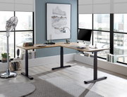 Moderner E-Eckschreibtisch in einem hellen Büro mit großen Fenstern, von vorne betrachtet. Der Schreibtisch hat eine Holzoberfläche und schwarze Metallbeine, ausgestattet mit einem Computer und Büchern.