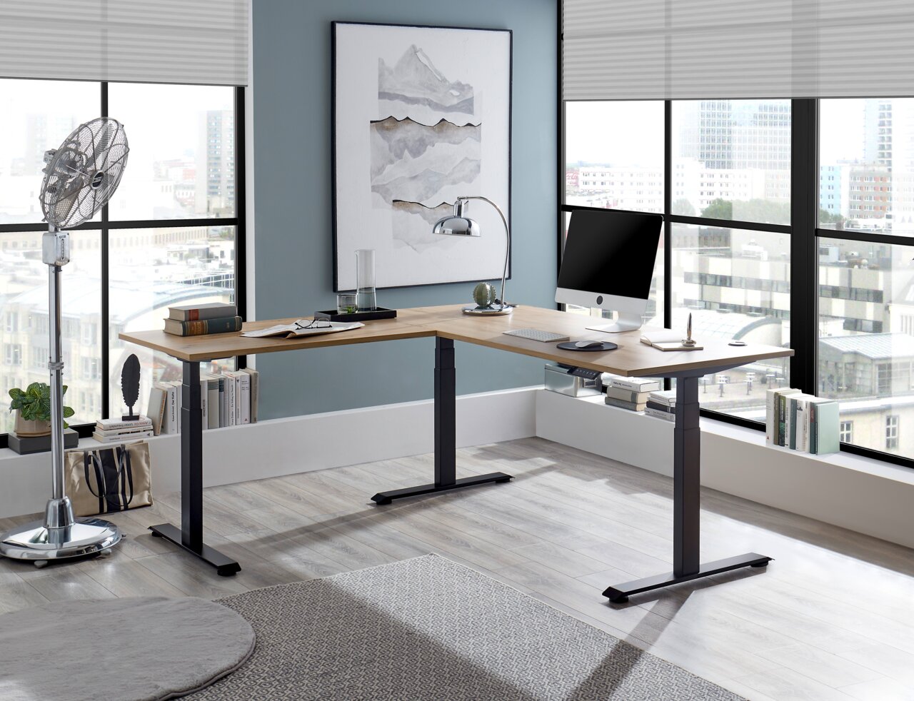 Moderner E-Eckschreibtisch in einem hellen Büro mit großen Fenstern, von vorne betrachtet. Der Schreibtisch hat eine Holzoberfläche und schwarze Metallbeine, ausgestattet mit einem Computer und Büchern.