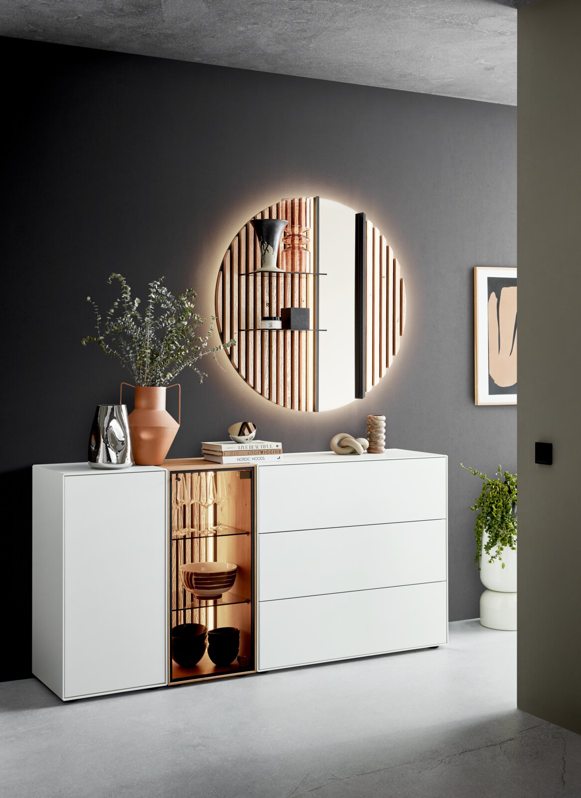 Modern wit dressoir met lades en een verlichte vitrinekast, versierd met vazen en boeken, tegen een donkere muur met een ronde spiegel. Aanzicht van voren.