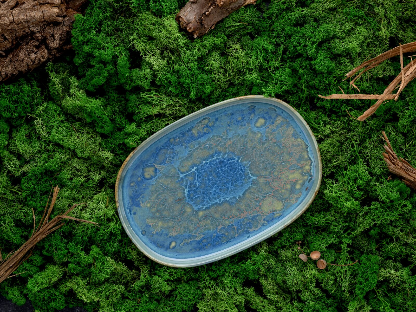 Ovale, handgefertigte Keramikschale in Blau mit reaktiver Glasur und hellen Sprenkeln, liegend auf grünem Moos mit Holzstücken, Draufsicht von oben