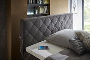 Seitliche Perspektive auf ein elegantes Boxspringbett mit grauem, gestepptem Kopfteil, grauer Bettwäsche und einem Tablet auf der Matratze.