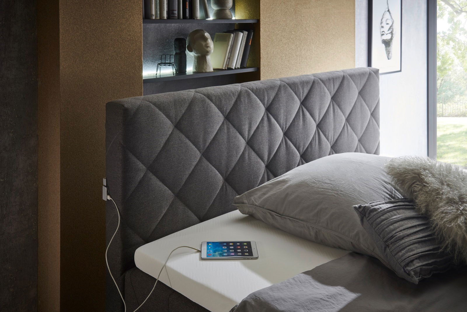 Seitliche Perspektive auf ein elegantes Boxspringbett mit grauem, gestepptem Kopfteil, grauer Bettwäsche und einem Tablet auf der Matratze.