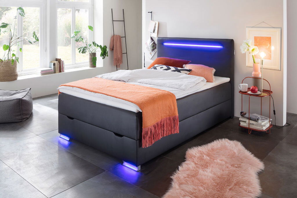 Z2 Boxbett LAS VEGAS Modernes Boxbett mit LED-Beleuchtung, seitliche Perspektive, dekoriert mit orangefarbenem Überwurf und Kissen, in einem hellen Raum mit großen Fenstern.