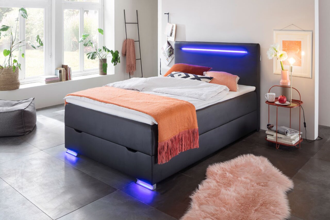 Modern boxbed met ledverlichting, gezien vanaf de zijkant, versierd met een oranje sprei en kussens, in een lichte kamer met grote ramen.