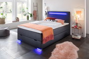 Modern boxbed met ledverlichting, gezien vanaf de zijkant, versierd met een oranje sprei en kussens, in een lichte kamer met grote ramen.