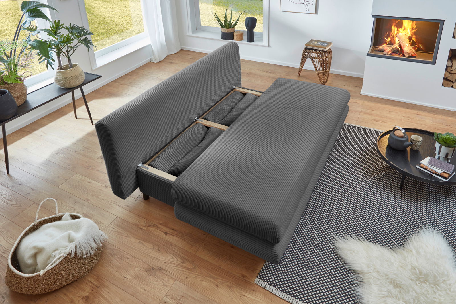 Graues Funktionssofa in einem modernen Wohnzimmer, von oben betrachtet, mit ausgeklappter Liegefläche, umgeben von Pflanzen, einem Kamin und einem Couchtisch.