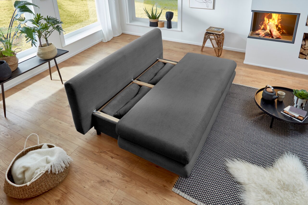 Graues Funktionssofa in einem modernen Wohnzimmer, von oben betrachtet, mit ausgeklappter Liegefläche, umgeben von Pflanzen, einem Kamin und einem Couchtisch.