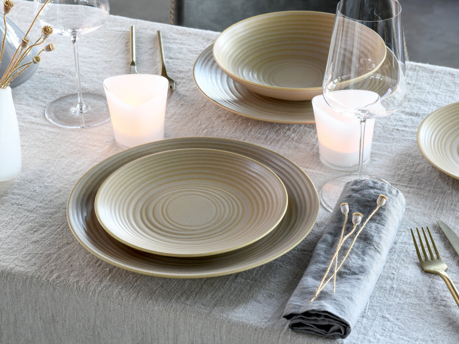 Schlichtes Keramikgeschirr-Set in Beige- und Grautönen mit Rillenstruktur auf gedecktem Tisch mit Weingläsern, Kerzen und grauer Leinenserviette, aufgenommen in leicht schräger Perspektive von oben