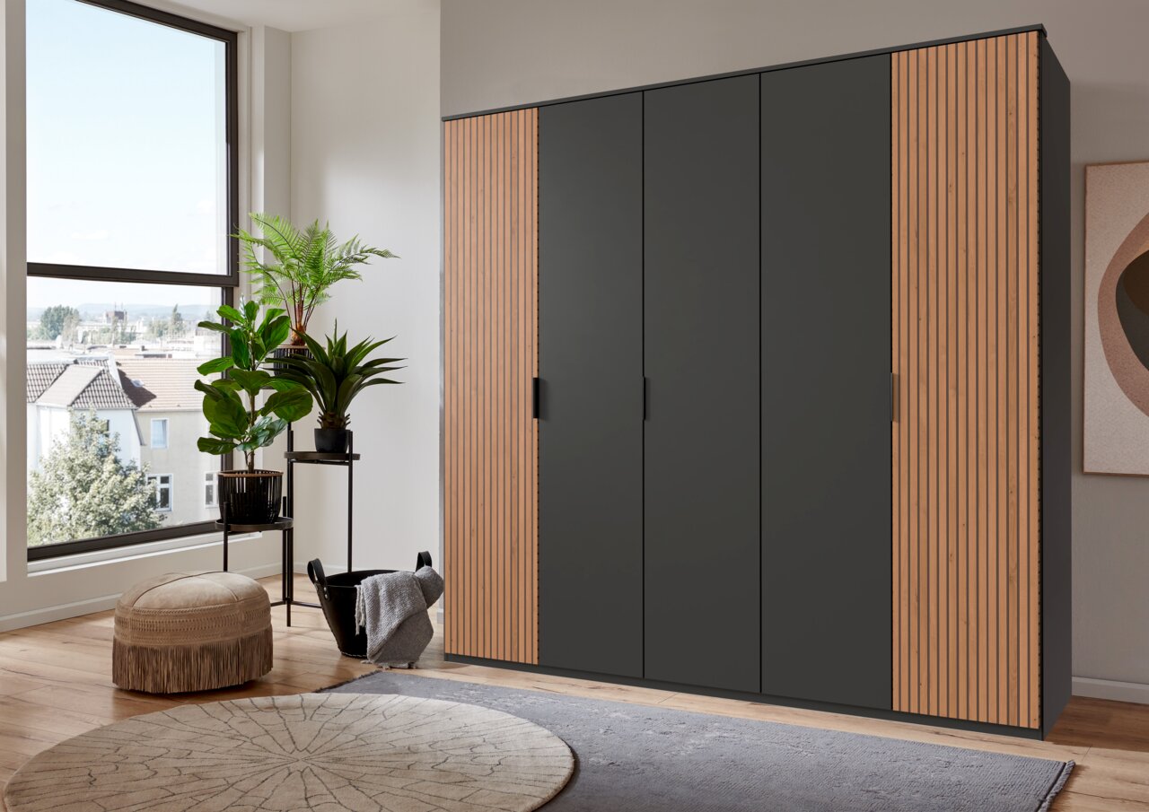 Moderner 5-türiger Drehtürenschrank in einem stilvollen Raum, frontal betrachtet. Der Schrank hat eine dunkle Oberfläche mit Holzdetails an den Seiten.
