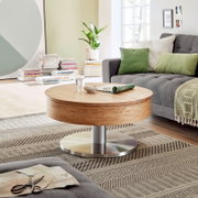 Moderne houten salontafel met rond tafelblad en metalen onderstel, in een gezellige woonkamer met een grijze bank en groene kussens, gezien vanuit de zijkant.