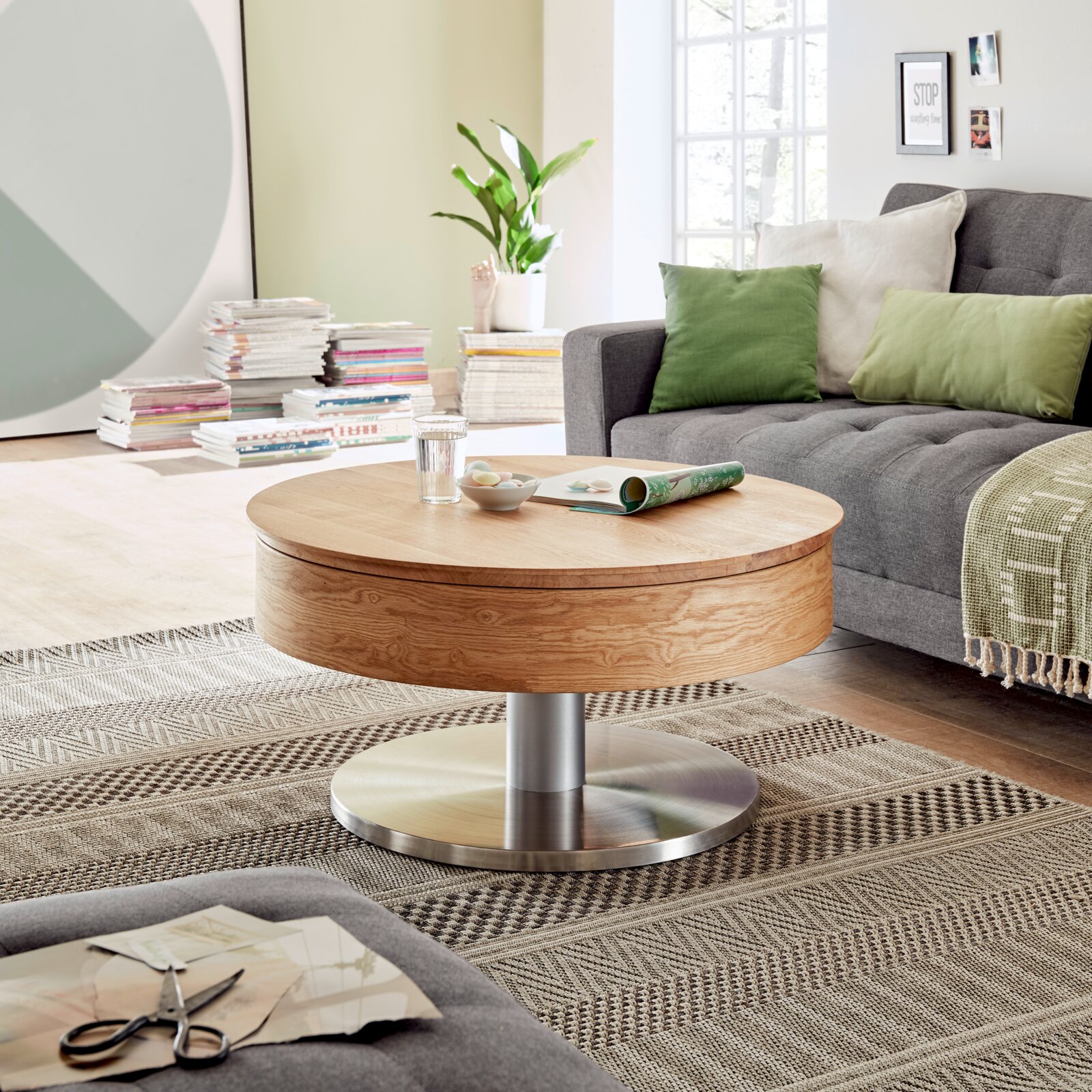 Moderne houten salontafel met rond tafelblad en metalen onderstel, in een gezellige woonkamer met een grijze bank en groene kussens, gezien vanuit de zijkant.