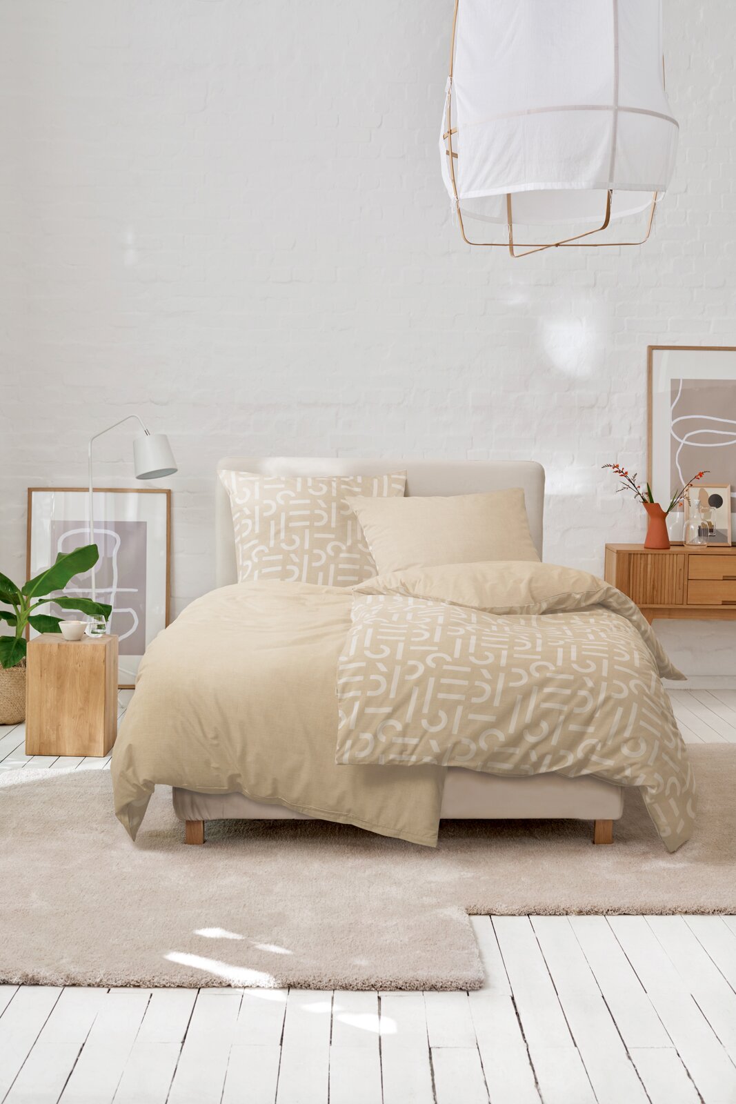 Beige Bettwäsche mit abstraktem Muster auf einem Doppelbett in hellem Schlafzimmer mit weißer Ziegelwand, Kissen und Deko, fotografiert in frontaler Perspektive leicht von oben.