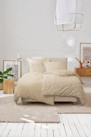 Beige Bettwäsche mit abstraktem Muster auf einem Doppelbett in hellem Schlafzimmer mit weißer Ziegelwand, Kissen und Deko, fotografiert in frontaler Perspektive leicht von oben.
