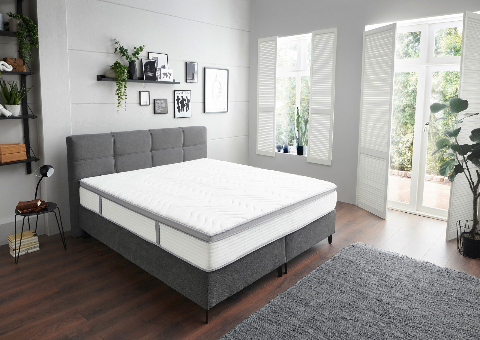 Modernes Boxspringbett in einem hellen, stilvoll eingerichteten Schlafzimmer mit grauem Kopfteil und weißer Matratze, seitliche Perspektive.