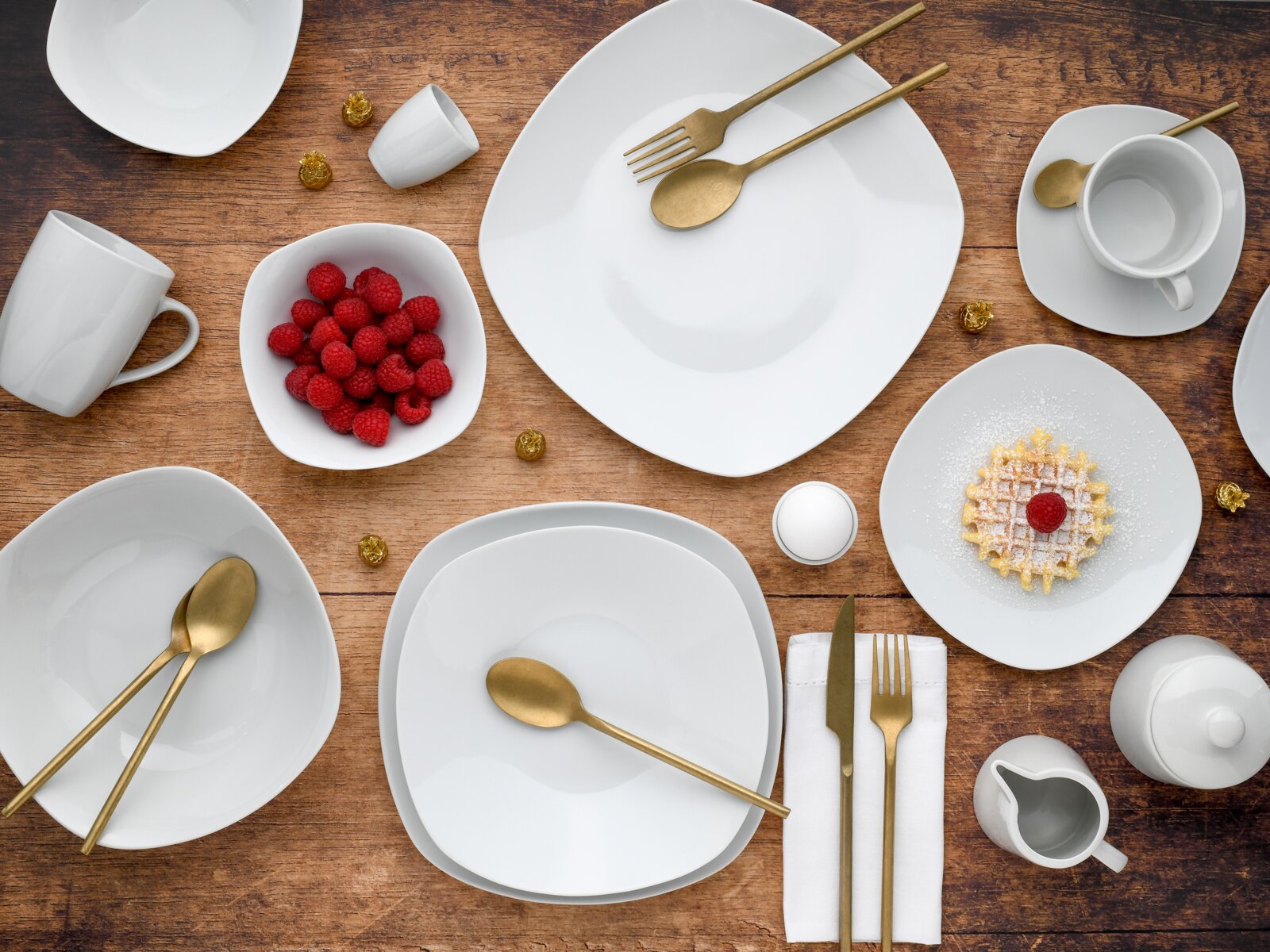 Draufsicht auf ein modernes weißes Geschirr-Set mit Tellern und Schalen auf Holztisch, dazu goldfarbenes Besteck, Tassen sowie Schale mit Himbeeren und ein kleines Waffel-Dessert.