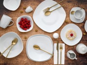 Draufsicht auf ein modernes weißes Geschirr-Set mit Tellern und Schalen auf Holztisch, dazu goldfarbenes Besteck, Tassen sowie Schale mit Himbeeren und ein kleines Waffel-Dessert.