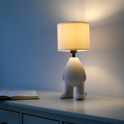 Een brandende tafellamp met een cilindervormige, crèmekleurige stoffen kap en een sculpturale voet in de vorm van een been, op een witte ladekast naast een open boek, gefotografeerd vanuit een licht zijdelings perspectief