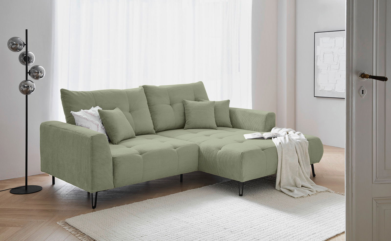 Grünes Ecksofa mit Ottomane rechts, dekoriert mit Kissen und Decke, in einem modernen Wohnzimmer mit hellem Holzboden und Stehlampe, Perspektive von vorne links.