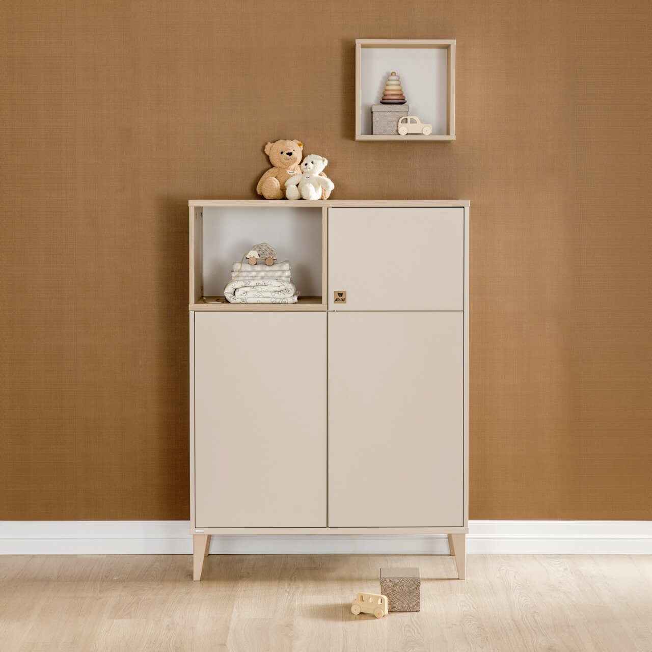 Beigefarbenes Highboard mit zwei Türen und einem offenen Fach, dekoriert mit Stofftieren und Babyzubehör, frontal fotografiert.