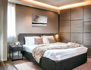 Modernes Schlafzimmer mit großem Doppelbett in grauem Polsterrahmen, weißer Bettwäsche und kuscheliger Decke, Nachttisch und Wandbild, aufgenommen aus schräger Frontperspektive.