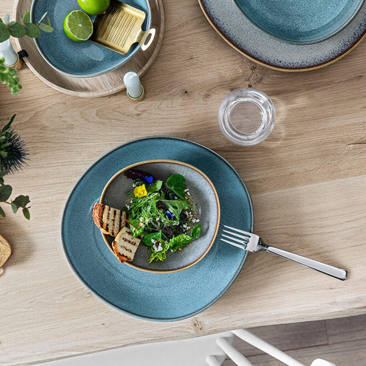 Like by Villeroy & Boch Schale flach LAVE BEIGE Vogelperspektive auf einen gedeckten Tisch mit einer Obstschale Lave 470ml beige, gefüllt mit Salat und gegrilltem Brot, umgeben von weiteren Tellern und Besteck.