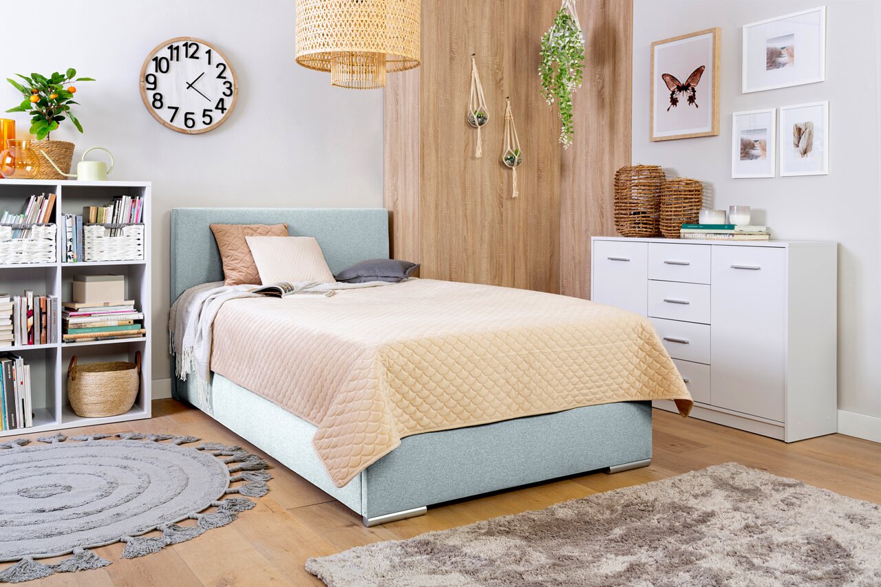 Gezellige slaapkamer met een boxbed met een lichtblauwe stoffen bekleding, versierd met kussens en een beige dekbed, omringd door moderne meubels en planten, gefotografeerd vanuit een zijdelingse hoek.