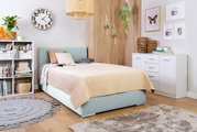 Gezellige slaapkamer met een boxbed met een lichtblauwe stoffen bekleding, versierd met kussens en een beige dekbed, omringd door moderne meubels en planten, gefotografeerd vanuit een zijdelingse hoek.
