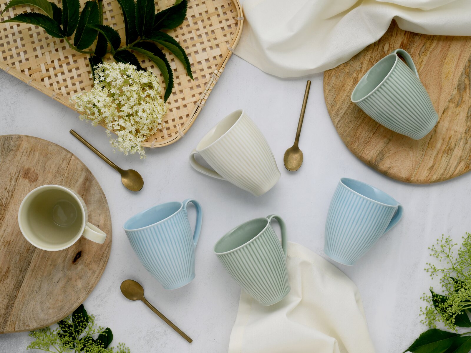 Draufsicht auf gerippte Keramik-Kaffeebecher in Pastellfarben (Blau, Grün, Creme) auf hellem Untergrund mit Holzbrettchen, goldenen Löffeln und Blumen dekorativ arrangiert