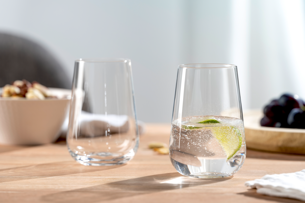 Leonardo Becher 6er-Set PALADINO Zwei elegante Gläser auf einem Holztisch, eines gefüllt mit sprudelndem Wasser und einer Limettenscheibe, im Hintergrund eine Schale mit Nüssen und Trauben, seitliche Perspektive.
