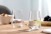 Twee elegante glazen op een houten tafel, waarvan één gevuld met bruisend water en een schijfje limoen, op de achtergrond een schaal met noten en druiven, zijaanzicht.
