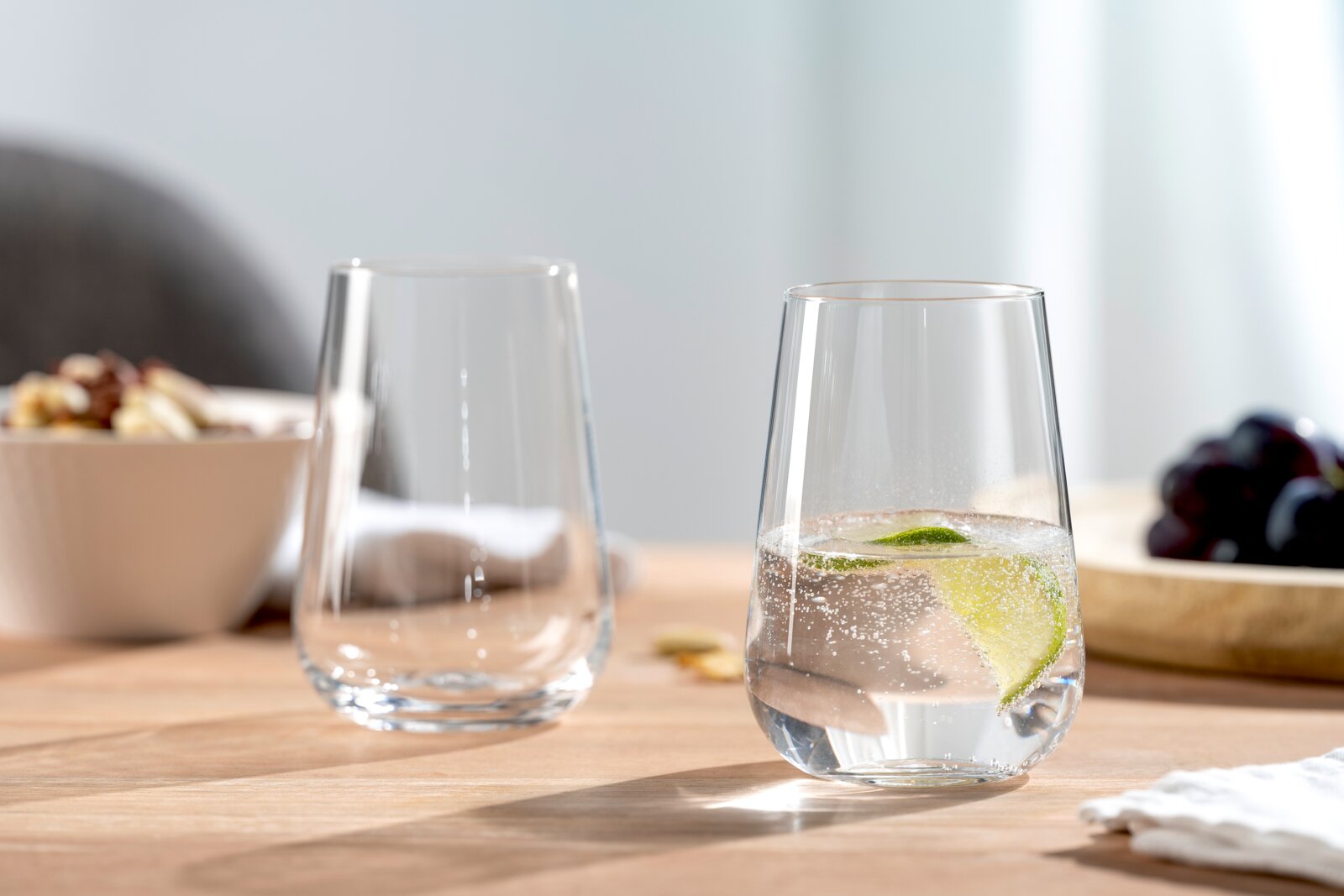 Twee elegante glazen op een houten tafel, waarvan één gevuld met bruisend water en een schijfje limoen, op de achtergrond een schaal met noten en druiven, zijaanzicht.