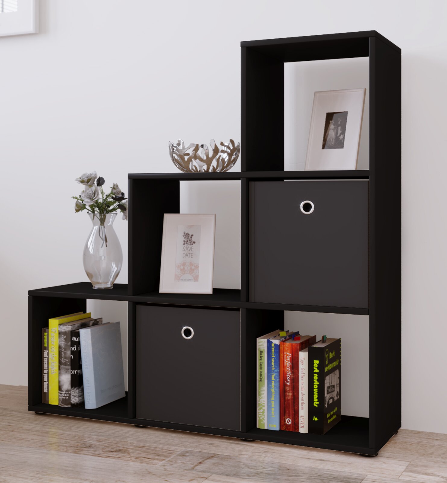 Modernes schwarzes Würfelregal/Standregal mit offenen Fächern und zwei Stoffboxen mit Metall-Öse, dekoriert mit Büchern, Vase und Schale; Perspektive: frontale Ansicht leicht von rechts.