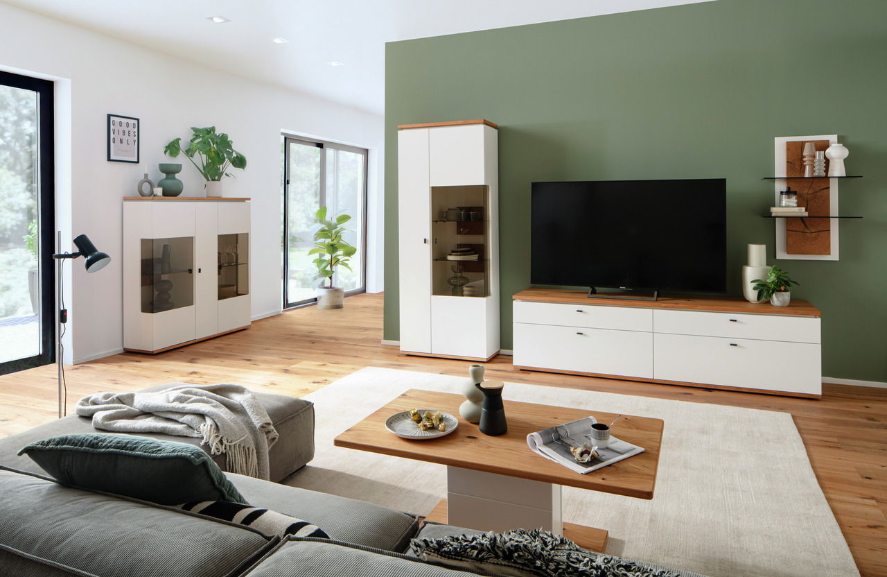 Moderne Wohnzimmer-Wandkombination MATADI in Weiß mit Holzdetails, bestehend aus einem TV-Schrank, einem hohen Schrank und einem Vitrinenschrank, aus der Perspektive eines gemütlichen Wohnzimmers mit Sofa und Couchtisch.