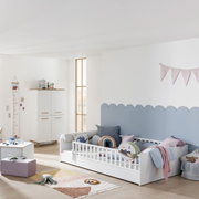 Kinderzimmer mit weißem Bodenbett und Rausfallschutz, dekoriert mit bunten Kissen und Decken, seitliche Perspektive