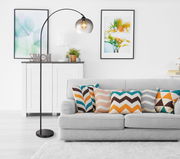 Moderne Stehleuchte NEWCASTLE mit gebogenem Arm und rauchigem Glasschirm, im Wohnzimmer neben einem grauen Sofa mit bunten Kissen, seitliche Perspektive.