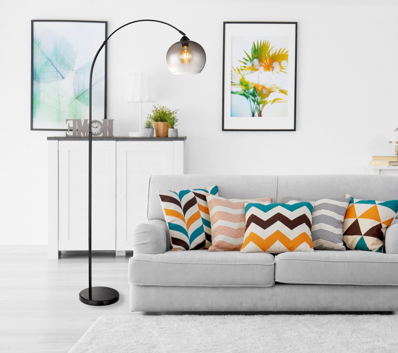 Moderne Stehleuchte NEWCASTLE mit gebogenem Arm und rauchigem Glasschirm, im Wohnzimmer neben einem grauen Sofa mit bunten Kissen, seitliche Perspektive.