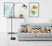 Moderne staande lamp NEWCASTLE met gebogen arm en rookkleurige glazen kap, in de woonkamer naast een grijze bank met kleurrijke kussens, zijaanzicht.