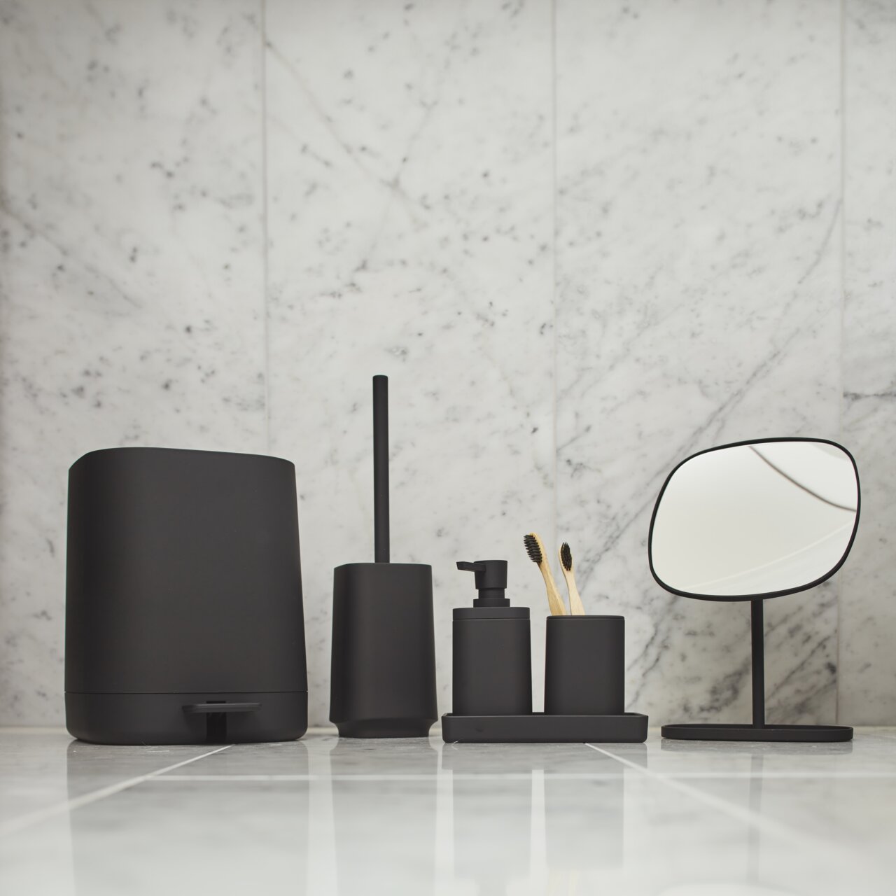 Modernes Badezimmer-Accessoire-Set in mattem Schwarz auf Fliesen vor Marmorwand: großer Behälter, WC-Bürste, Seifenspender und Becher mit Zahnbürsten auf Ablage sowie kleiner Standspiegel, frontal auf Augenhöhe fotografiert.