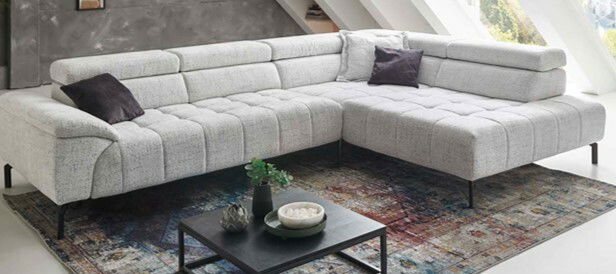 Beldomo Style Ecksofa VEGAS Eckgarnitur in hellem Stoff mit schwarzen Beinen, von schräg vorne fotografiert, auf einem gemusterten Teppich stehend.