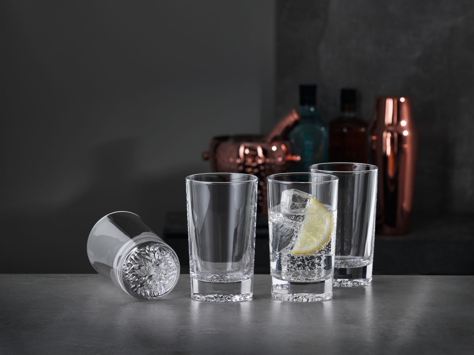 Drei aufrecht stehende Softdrinkgläser und ein liegendes Glas auf einer dunklen Oberfläche, mit einem Glas gefüllt mit einem Getränk, Eiswürfeln und einer Zitronenscheibe, im Hintergrund Barzubehör.