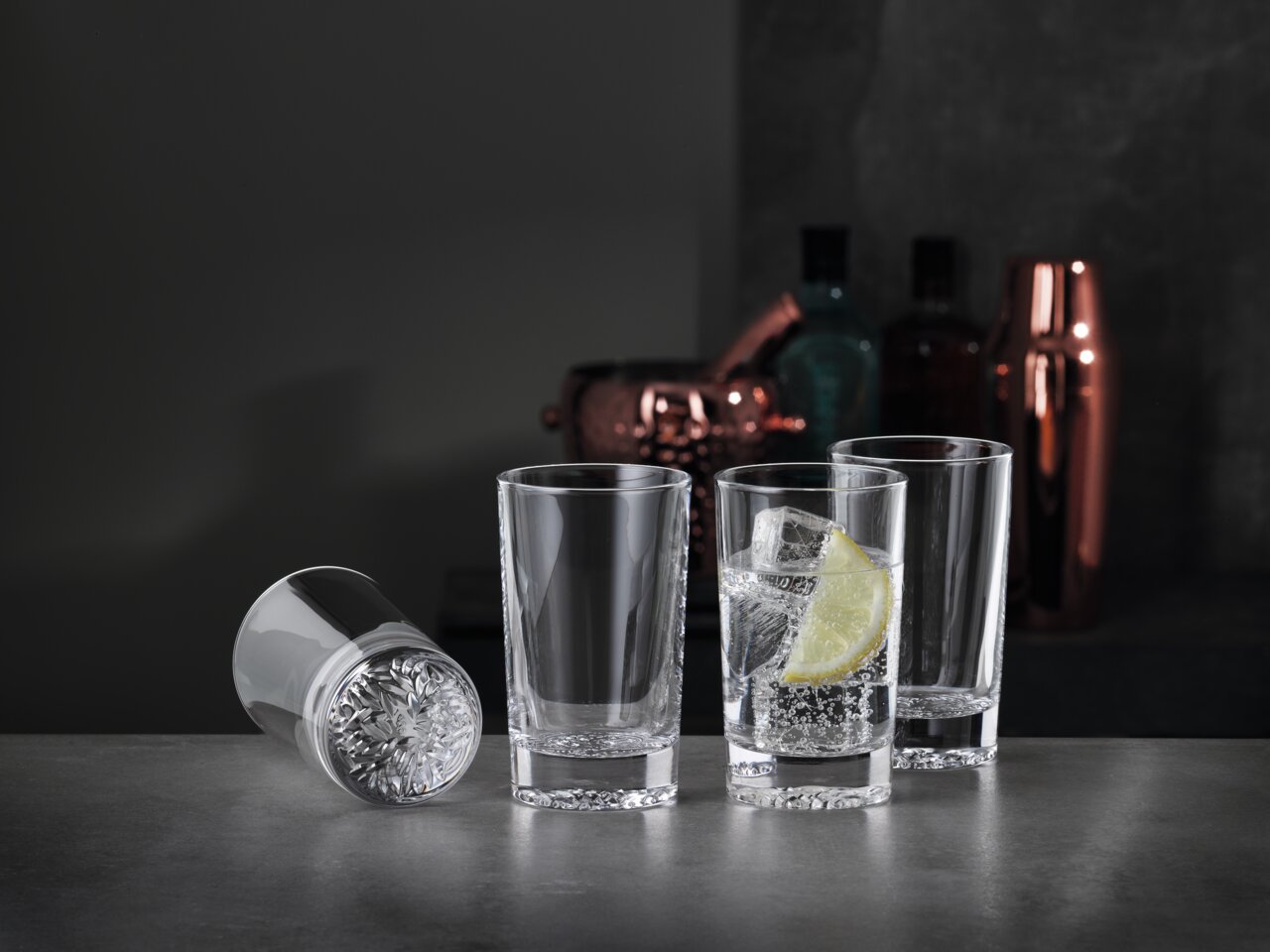 Drei aufrecht stehende Softdrinkgläser und ein liegendes Glas auf einer dunklen Oberfläche, mit einem Glas gefüllt mit einem Getränk, Eiswürfeln und einer Zitronenscheibe, im Hintergrund Barzubehör.