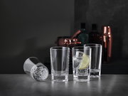 Drei aufrecht stehende Softdrinkgläser und ein liegendes Glas auf einer dunklen Oberfläche, mit einem Glas gefüllt mit einem Getränk, Eiswürfeln und einer Zitronenscheibe, im Hintergrund Barzubehör.