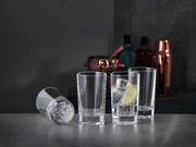 Drei aufrecht stehende Softdrinkgläser und ein liegendes Glas auf einer dunklen Oberfläche, mit einem Glas gefüllt mit einem Getränk, Eiswürfeln und einer Zitronenscheibe, im Hintergrund Barzubehör.